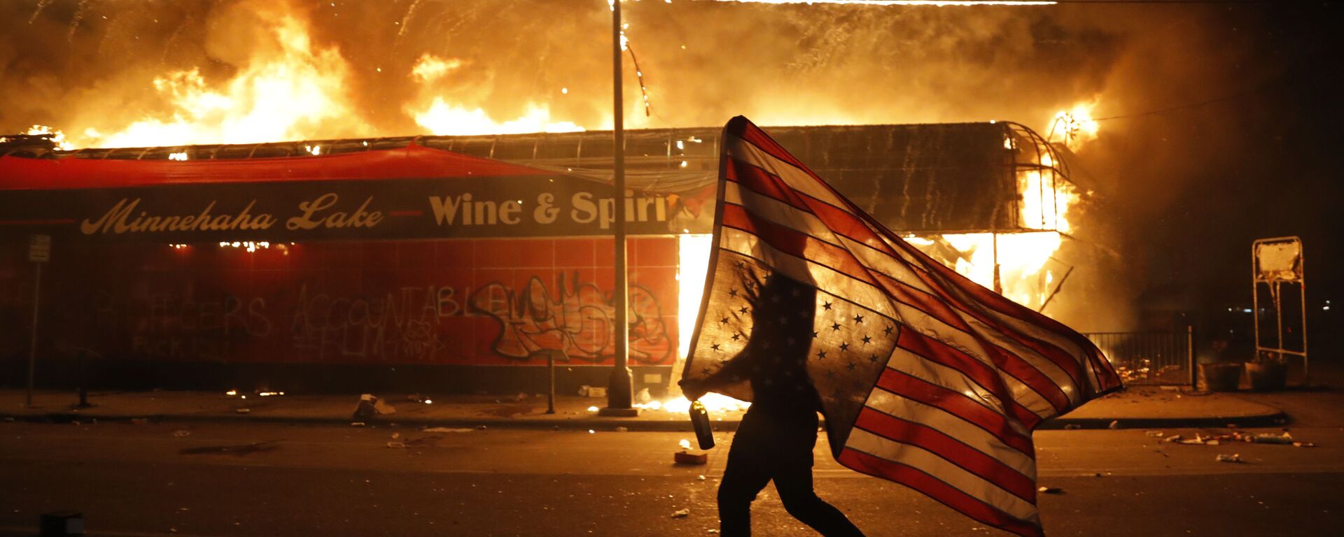 Un manifestante porta la bandera de EEUU boca abajo como símbolo de peligro extremo que vive el país frente un edificio en llamas en Minneapolis (estado de Minnesota), durante las protestas causadas por la muerte de George Floyd, el 28 de mayo del 2020 - Sputnik Mundo, 1920, 08.08.2022
