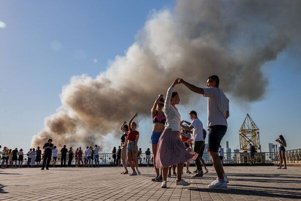 Unas parejas bailan samba con el humo de un almacén pirotécnico en llamas en Moscú de fondo. - Sputnik Mundo