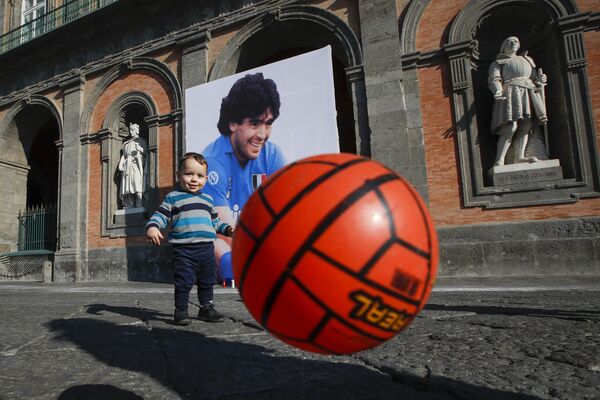 Andrea Cangiano, de 16 meses, juega con un balón frente a una imagen de Maradona delante del Palacio Real de Nápoles (Italia) el 27 de noviembre de 2020. El crac argentino jugó por el Nápoles entre los años de 1984 y 1992. - Sputnik Mundo
