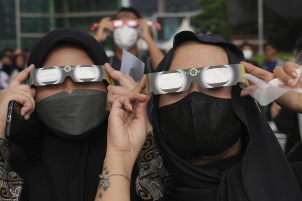Los pocos afortunados que se encuentren en la trayectoria del eclipse se verán sumidos en la oscuridad total u observarán un anillo de fuego cuando el Sol asome por detrás de la Luna.En la foto: unas mujeres indonesias usan gafas protectoras para ver el eclipse en Yakarta, Indonesia. - Sputnik Mundo