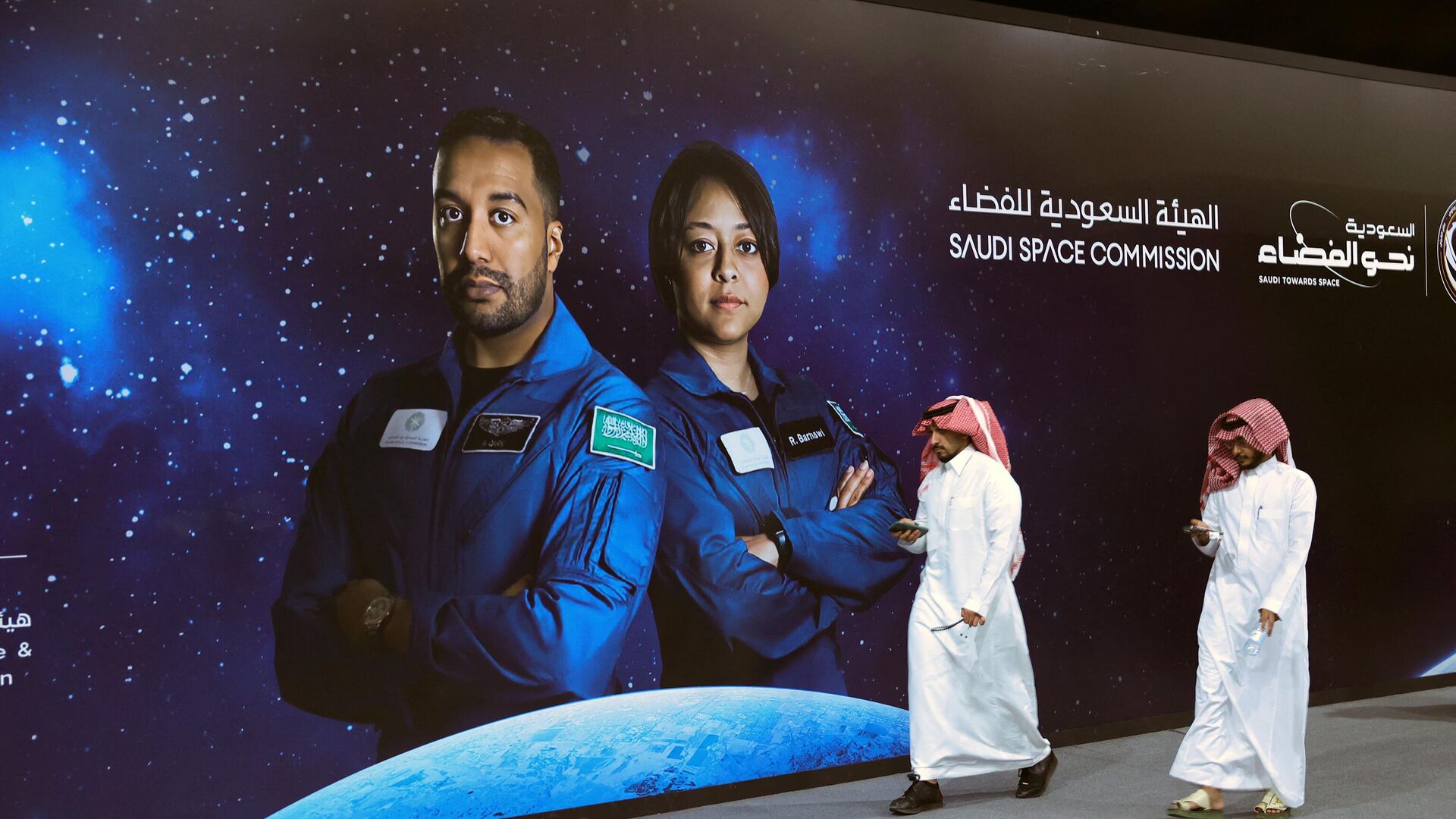 Un grupo de personas pasa junto a un cartel con la foto de los astronautas de la Comisión Espacial Saudita Ali Al-Qarni (izq.) y Rayyanah Barnawi, de la Misión Axiom 2, en una fiesta organizada por el gobierno en Riad el 21 de mayo de 2023.  - Sputnik Mundo, 1920, 22.05.2023