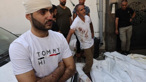 Un palestino herido junto a otras personas junto a los cuerpos de las víctimas de los ataques aéreos israelíes frente al depósito de cadáveres del hospital Al Shifa en la ciudad de Gaza el 12 de octubre de 2023. - Sputnik Mundo